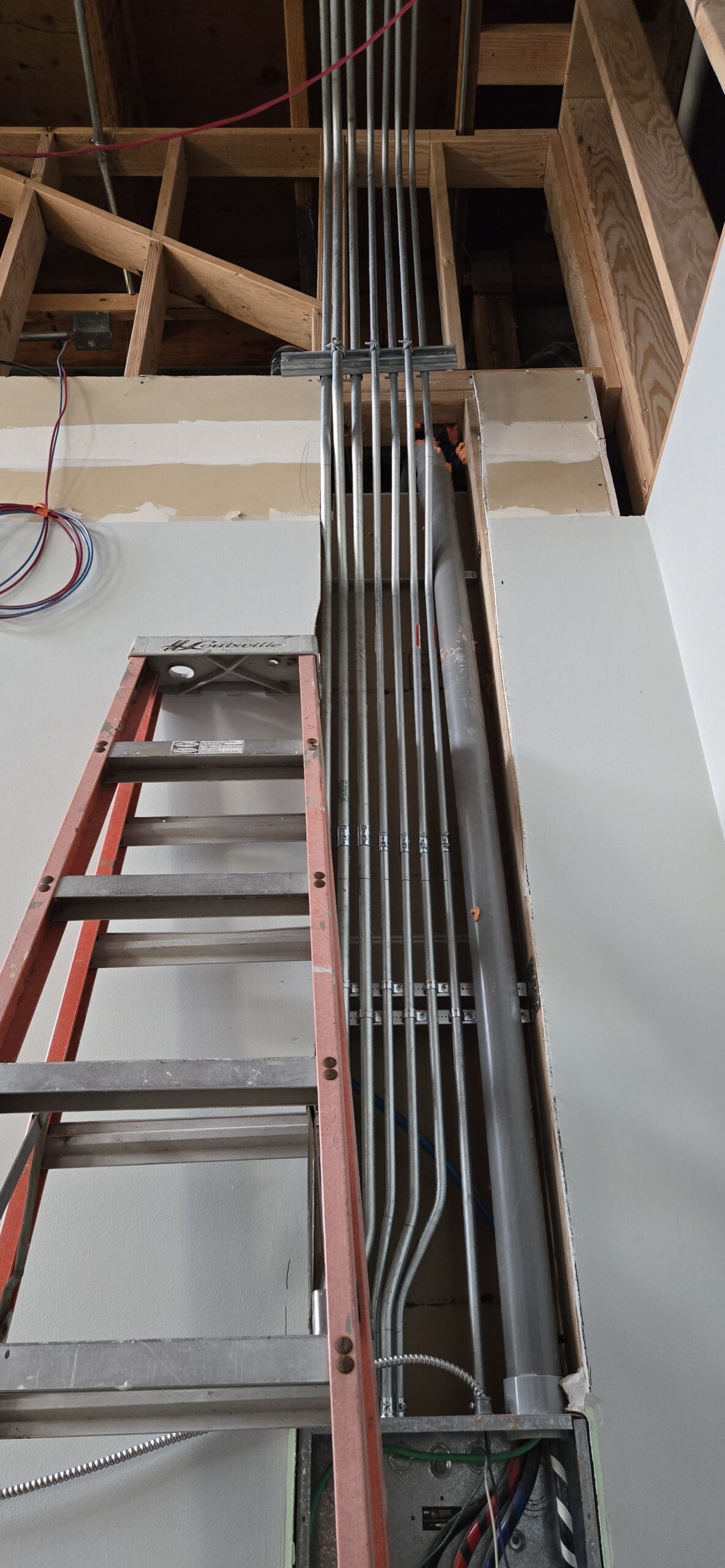 A red ladder is positioned against a wall, leading to a partially exposed ceiling with multiple metal conduits and some visible wires.