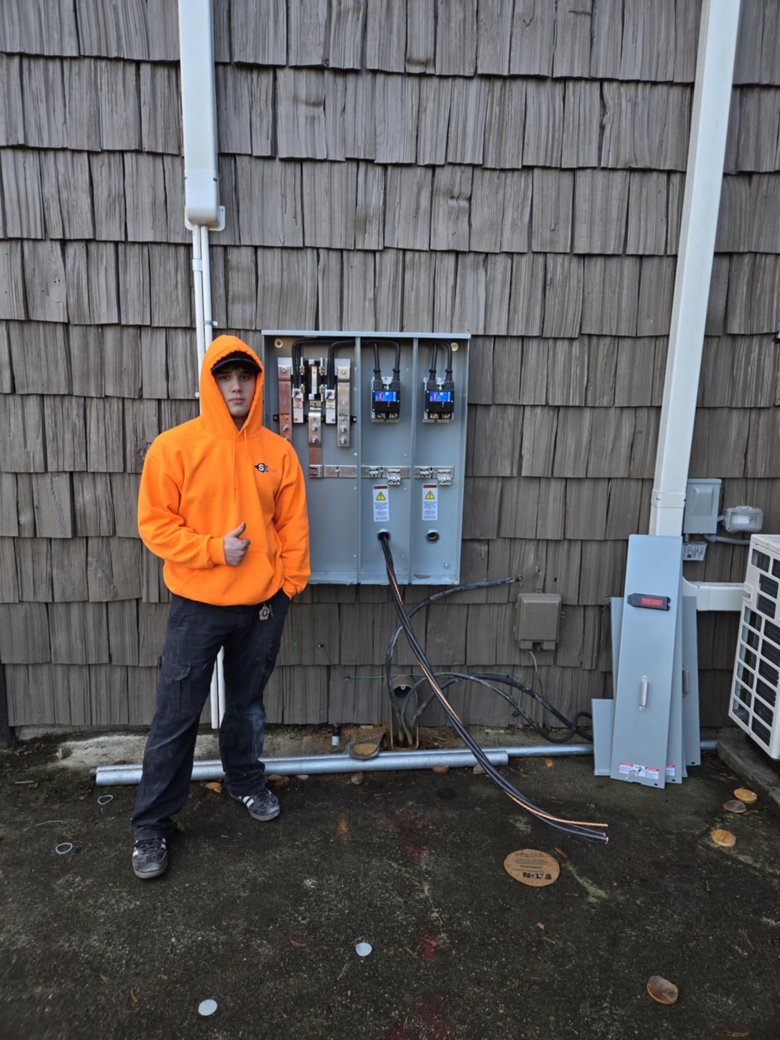 Person in an orange hoodie stands next to an open electrical panel with exposed cables on a textured wall.