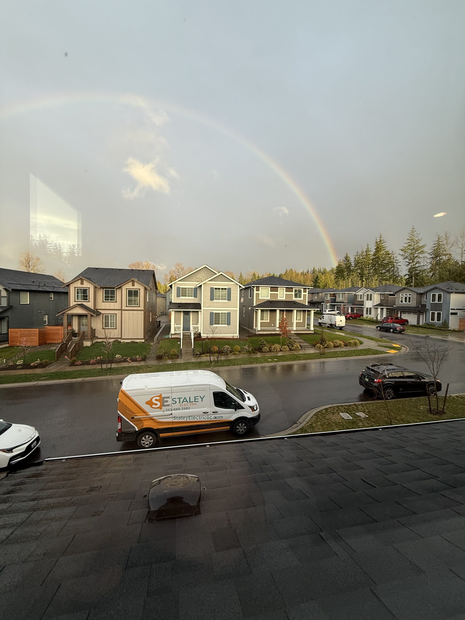 A residential street with houses and parked cars. A rainbow arches over the scene. A white van with company branding is parked on the road.