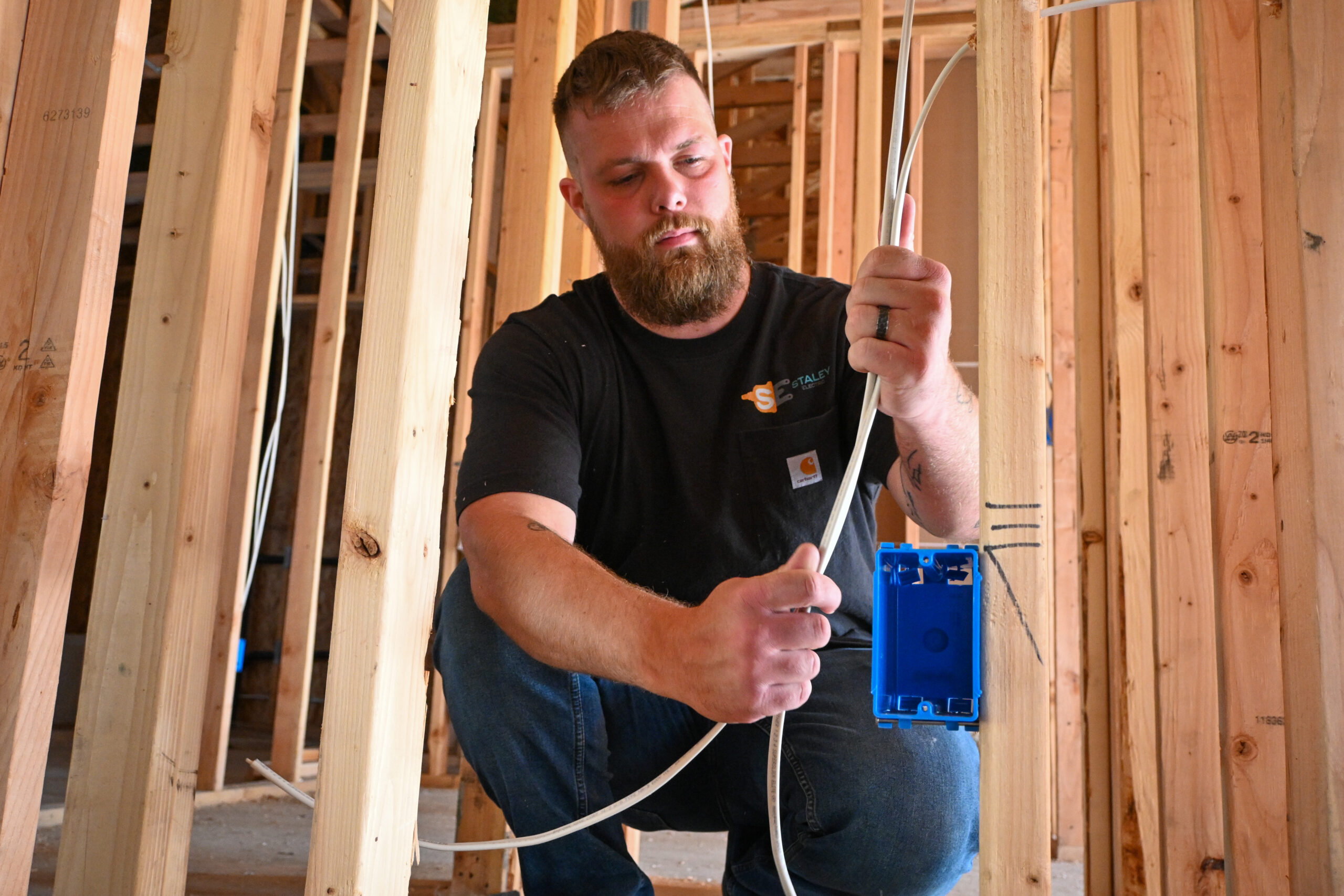 A person installs electrical wiring in a wooden frame structure, holding a blue electrical box.