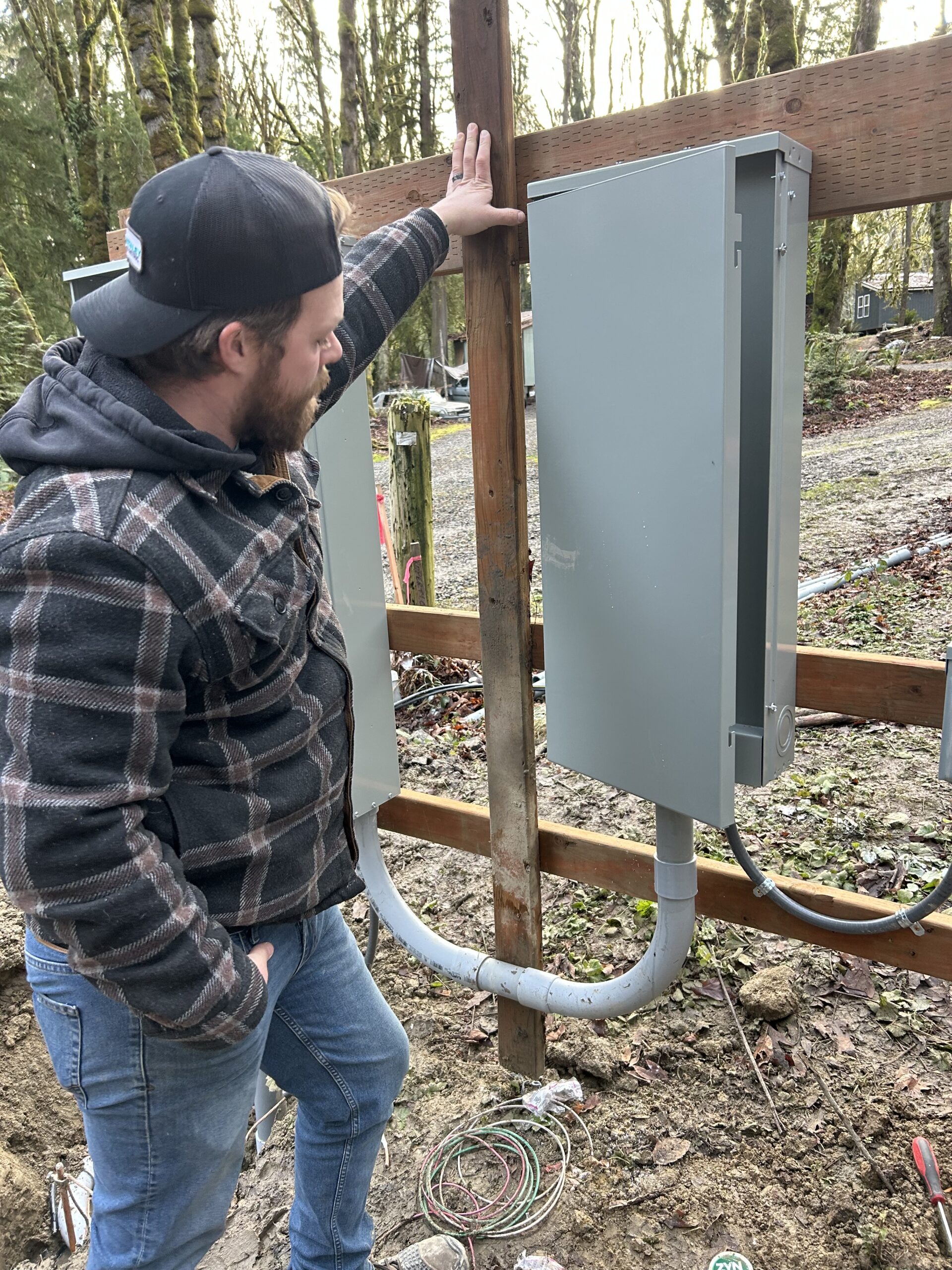 A man in a plaid jacket and cap stands outdoors, looking at two gray electrical panels mounted on a wooden board. The area is surrounded by trees and unpaved ground.
