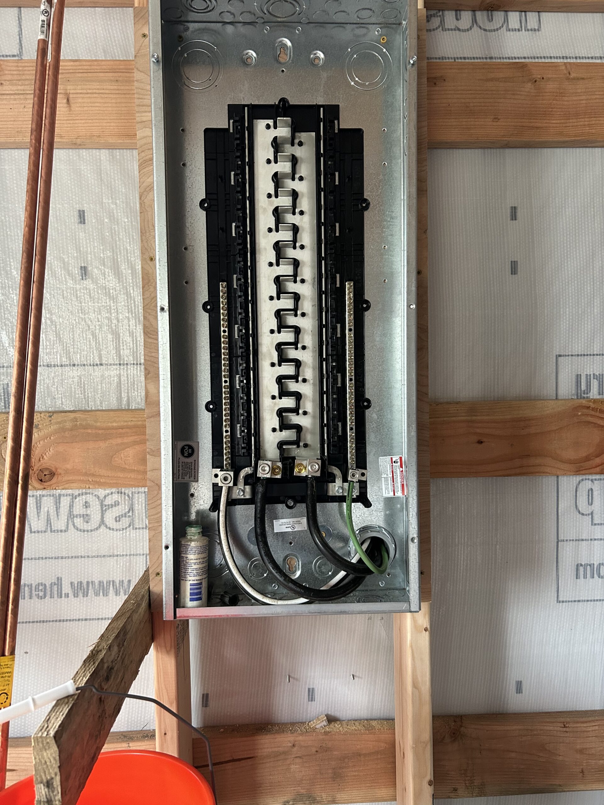 Open electrical panel on wall with visible circuit breakers and wiring. A small bottle is attached at the bottom left. Background consists of wooden beams and building wrap.