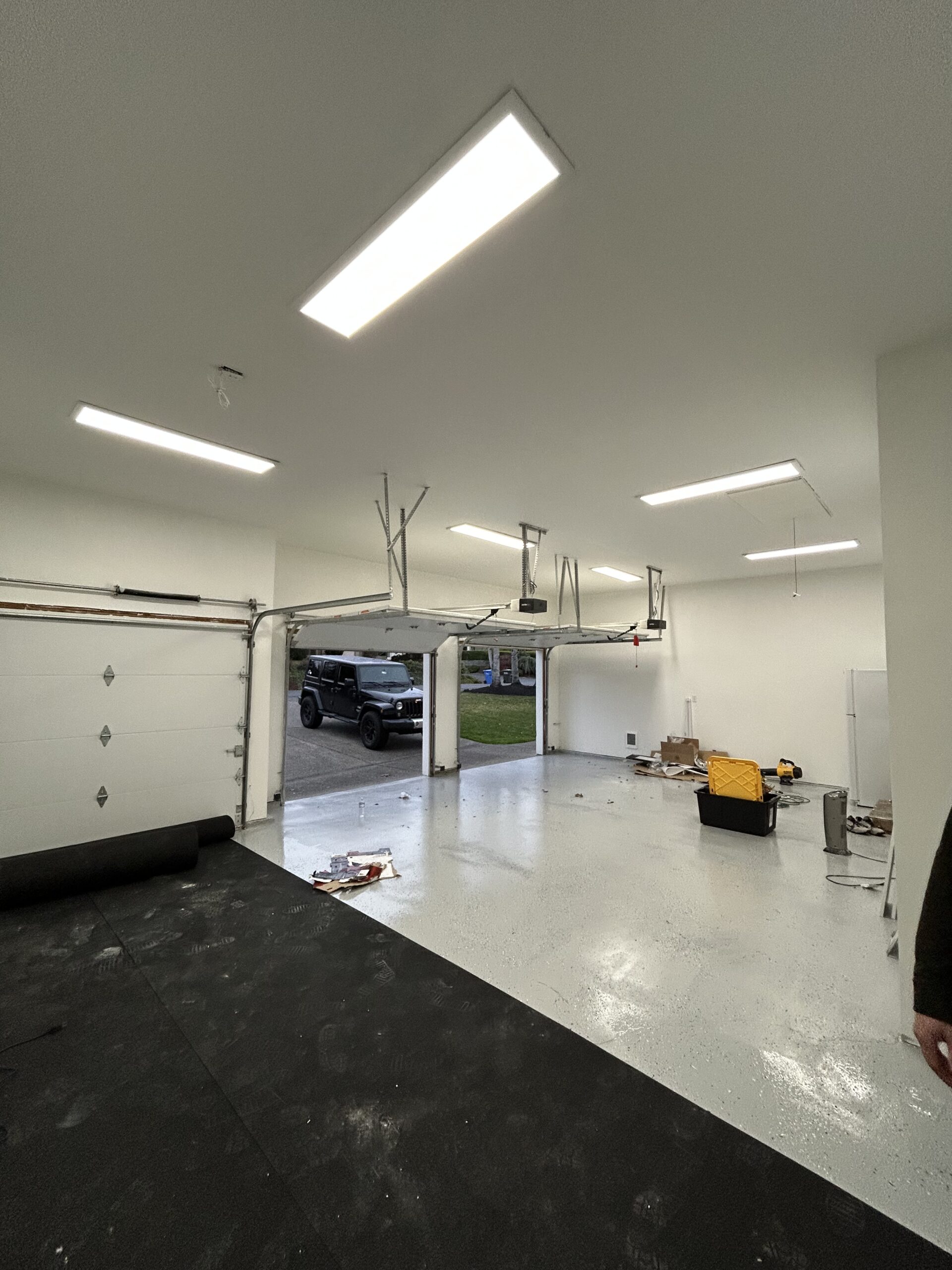 Garage interior with open double doors showing a parked truck outside. Floor partly covered with black mats; various tools and boxes scattered around. Overhead lights are on.