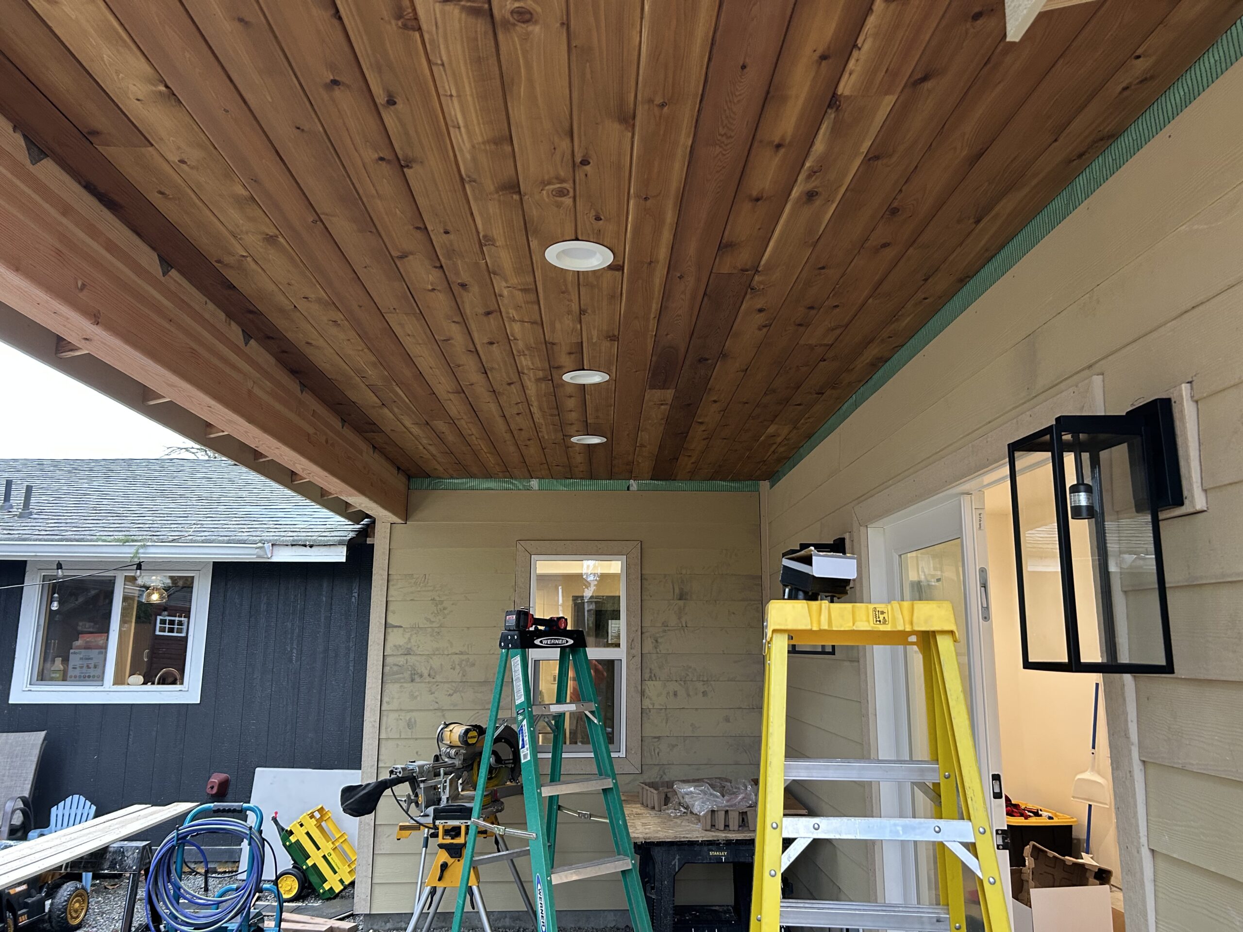 Construction site showing a covered porch with a wood ceiling, ladders, tools, and building materials.